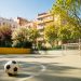 Pista de fútbol de barrio vacía en un patio de Madrid al atardecer, con porterías desgastadas, un balón sobre las líneas pintadas y bancos de madera junto a fachadas antiguas con balcones